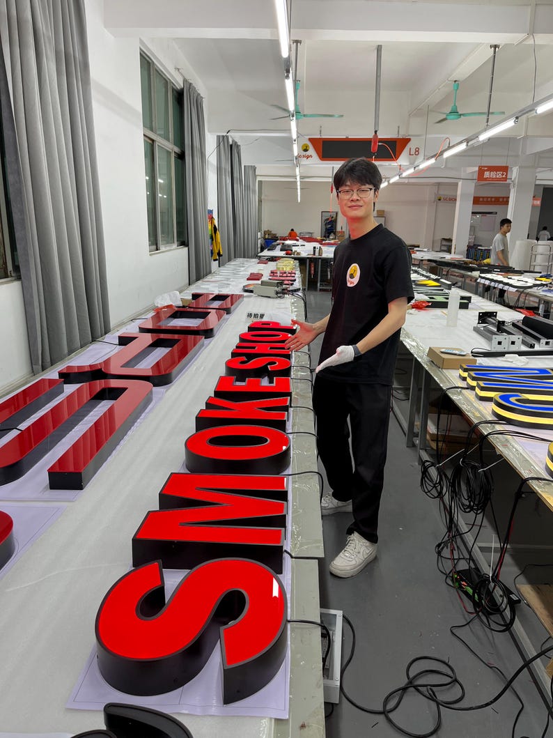 May include: Large, illuminated red and black letters spelling "SMOKE" are displayed on a table in a workshop. The letters are three-dimensional and appear to be made of acrylic or a similar material. A person is standing next to the letters.
