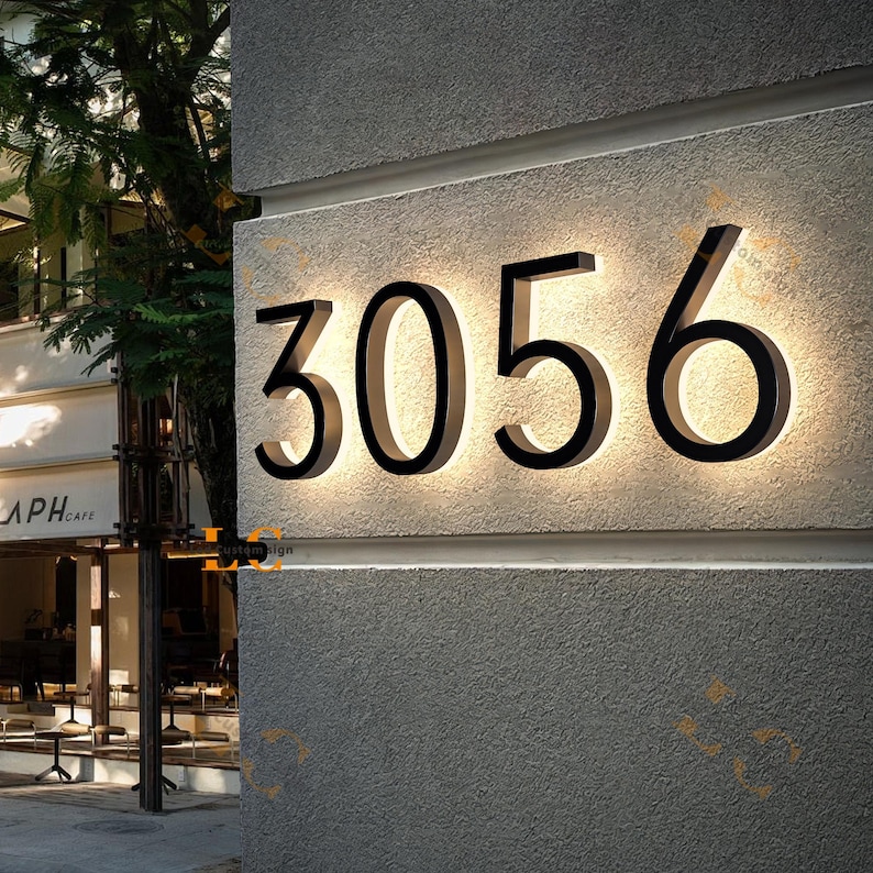 Puede incluir: N&uacute;meros de casa negros iluminados 3056 montados en una pared gris texturizada. Los n&uacute;meros est&aacute;n delineados con una luz brillante, creando un aspecto moderno y elegante. El fondo incluye un caf&eacute; y &aacute;rboles.