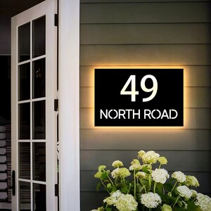 May include: A black illuminated address sign displaying the number "49" and the words "NORTH ROAD" in white. The sign is mounted on a gray wall with a warm, yellow glow around the edges. White flowers are in the foreground.