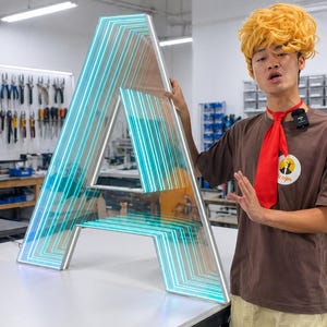 May include: Large illuminated letter "A" with a neon blue and white glow. The letter is made of clear acrylic with a layered design. The sign is on a white surface. The sign is likely for a business or event.