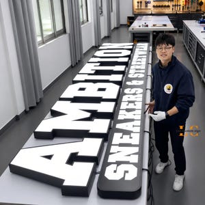 May include: A large, custom-made, illuminated channel letter sign featuring the text AMBITION SNEAKERS & STREETWEAR in bold, white, block-style lettering against a black background, displayed on a long, flat, white work surface in a workshop.