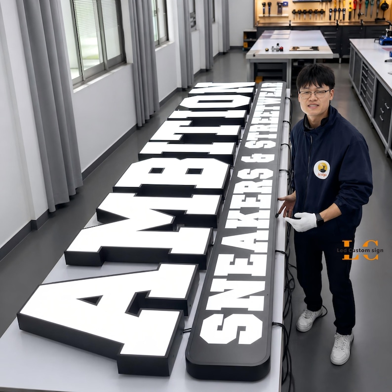 May include: A large, custom-made, illuminated channel letter sign featuring the text AMBITION SNEAKERS & STREETWEAR in bold, white, block-style lettering against a black background, displayed on a long, flat, white work surface in a workshop.