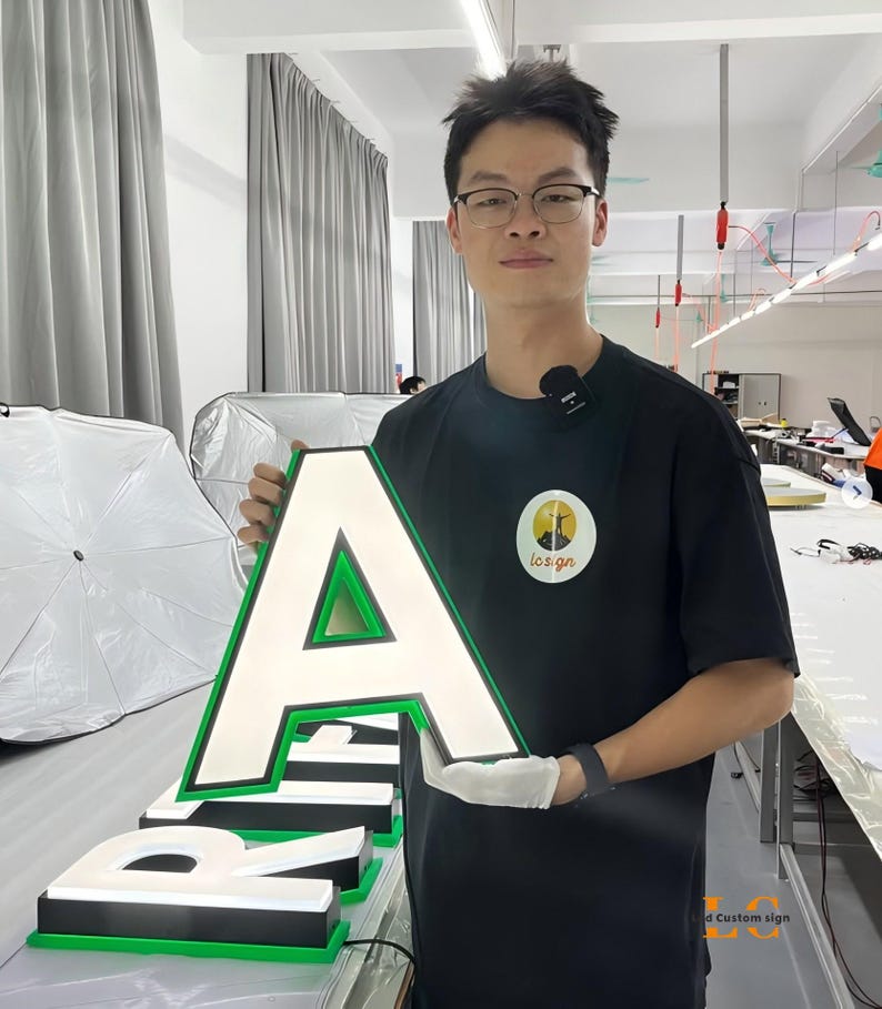 May include: A large, three-dimensional letter A sign featuring a white illuminated face, a black border, and a vibrant green trim. The letter is constructed with a thick profile and is designed for high-visibility signage applications.