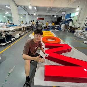 Peut inclure: Une grande lettre "N" rouge et une lettre "O" orange illuminées sont affichées sur une surface blanche dans un atelier. Les lettres font partie d'une enseigne plus grande. L'image montre également une personne en chemise marron et short noir.