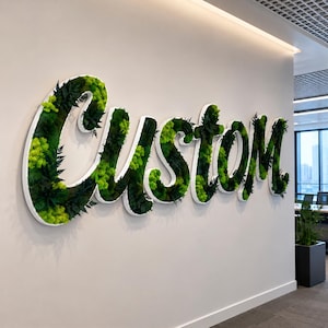 May include: A large wall-mounted sign spelling the word Custom in cursive script. The letters feature a white border and are filled with preserved green moss and dark green fern foliage, creating a textured, three-dimensional botanical display.