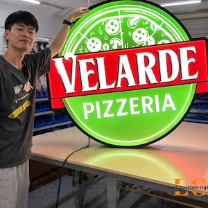 May include: A round, illuminated sign for a pizzeria. The sign features a green pizza graphic with white mushrooms and pepperoni. The text "VELARDE PIZZERIA" is displayed in bold white letters on a red banner. The sign is on a table.