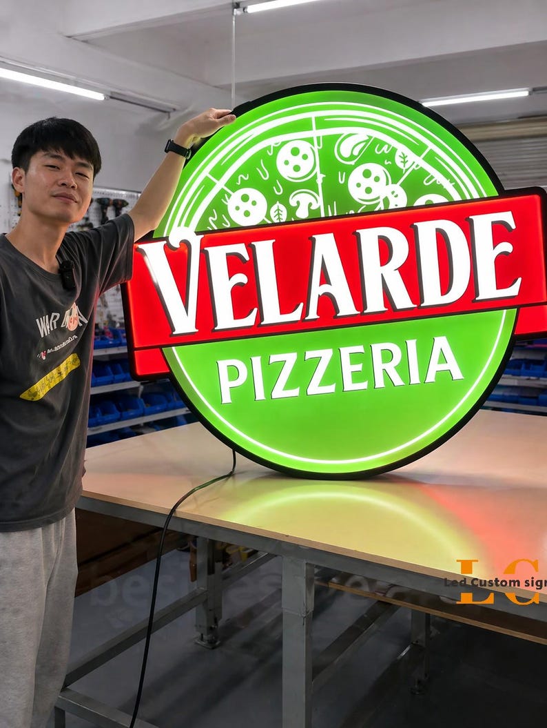 May include: A round, illuminated sign for a pizzeria. The sign features a green pizza graphic with white mushrooms and pepperoni. The text "VELARDE PIZZERIA" is displayed in bold white letters on a red banner. The sign is on a table.