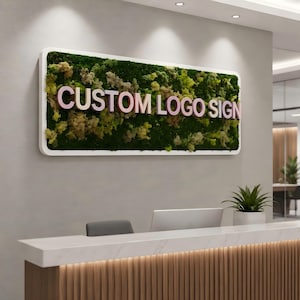 May include: A rectangular wall sign featuring a white frame filled with preserved green moss and light-colored three-dimensional lettering that reads CUSTOM LOGO SIGN. The sign is mounted on a wall above a reception desk in an office setting.