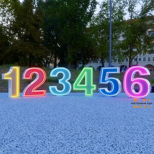 K&ouml;nnte beinhalten: Gro&szlig;e, beleuchtete Zahlen 1 bis 6, jede mit einer anderen Neonfarbe: Gelb, Rot, Hellblau, Gr&uuml;n, Dunkelblau und Pink. Die Zahlen werden im Freien auf einer strukturierten Oberfl&auml;che angezeigt.