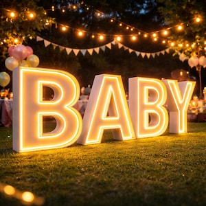 Puede incluir: Grandes letras "BABY" iluminadas con un brillo amarillo cálido, colocadas sobre césped. Las letras son blancas con un contorno amarillo, creando un ambiente festivo. Luces de cadena y globos son visibles en el fondo, sugiriendo una celebración.