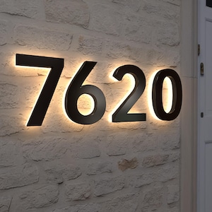 May include: Black illuminated house numbers, displaying "7620", mounted on a textured, light-colored stone wall. The numbers are backlit with a warm, inviting glow, creating a modern and stylish look for home exteriors.