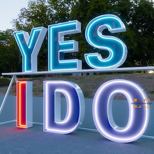 K&ouml;nnte beinhalten: Gro&szlig;es beleuchtetes Schild mit den Worten "YES I DO" in Wei&szlig; mit neonblauen und roten Akzenten. Die Buchstaben sind gro&szlig; und stehen auf einer Oberfl&auml;che, perfekt f&uuml;r eine Hochzeit oder Veranstaltung. Das Schild befindet sich im Freien mit B&auml;umen im Hintergrund.