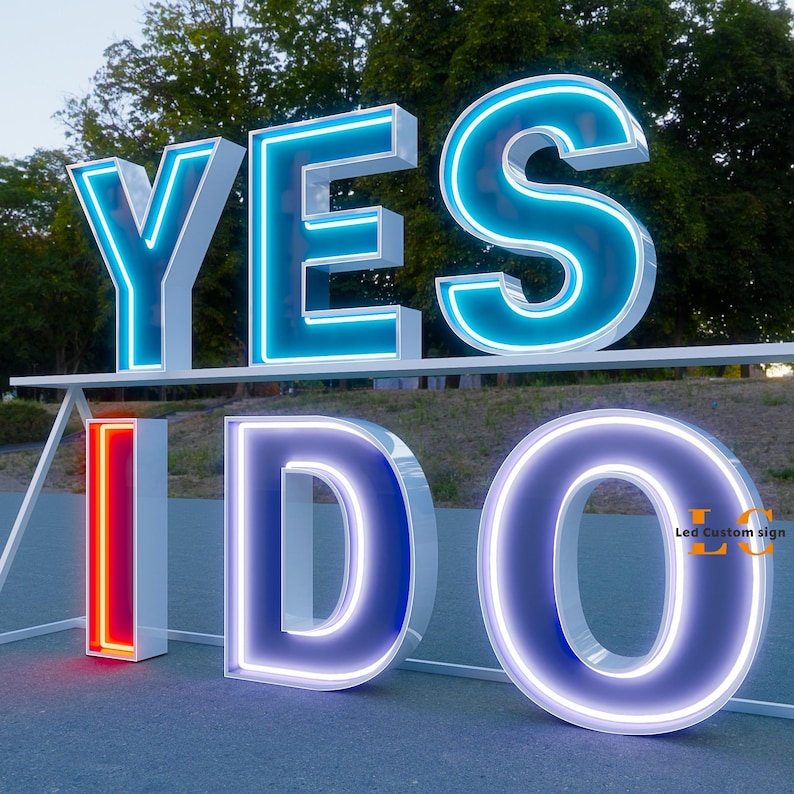 K&ouml;nnte beinhalten: Gro&szlig;es beleuchtetes Schild mit den Worten "YES I DO" in Wei&szlig; mit neonblauen und roten Akzenten. Die Buchstaben sind gro&szlig; und stehen auf einer Oberfl&auml;che, perfekt f&uuml;r eine Hochzeit oder Veranstaltung. Das Schild befindet sich im Freien mit B&auml;umen im Hintergrund.