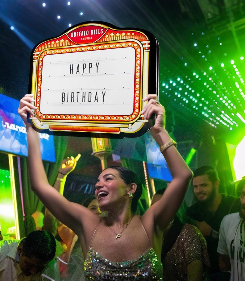 May include: A decorative sign with the text "HAPPY BIRTHDAY" in a marquee style. The sign has a red and gold border with the words "BUFFALO BILLS" at the top. The sign is held up by a person in a sparkly dress.