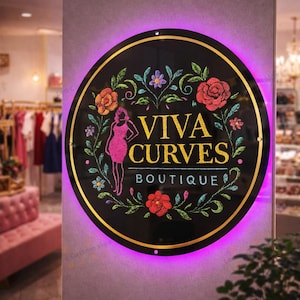 May include: A round black sign with gold trim and the words "VIVA CURVES BOUTIQUE" in gold lettering. The sign features a pink silhouette of a woman and is surrounded by colorful floral designs. The sign is illuminated with a purple glow.