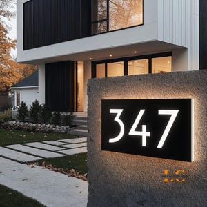 May include: Black rectangular address sign with the number 347 in white, illuminated against a stone pillar. The sign is mounted outdoors in front of a modern house with a white exterior and black accents. The sign is lit with a warm glow.