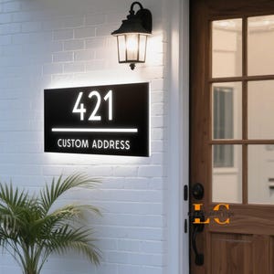 May include: A black custom address sign with the number "421" in white, illuminated by a white light. The words "CUSTOM ADDRESS" are below the number. The sign is mounted on a white brick wall, with a black lantern above.