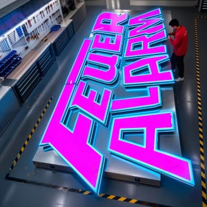 May include: Large, illuminated sign spelling out "FEUER ALARM" in bright pink letters with a blue outline. The sign is in a workshop setting, with a person working on it. The letters are large and three-dimensional.
