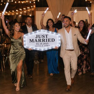 May include: A white, hexagonal, LED-lit marquee sign featuring the black text 'JUST MARRIED', the date '05.18.2025', and the names 'OLIVIA & JAMES'. The sign is illuminated by a series of small, bright bulbs along its perimeter, held by two people.