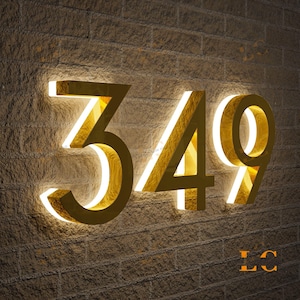 May include: Illuminated gold-colored house numbers 349 mounted on a textured brick wall. The numbers have a bright white internal glow, creating a striking contrast against the warm-toned wall. The numbers are in a modern font.