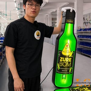 May include: A large, illuminated green bottle-shaped sign with the text "ZURE BOM SHOTJE" and "Veere de Kesbeke" is held by a person. The sign is black-bordered and has a matching base. The person is wearing a black t-shirt.