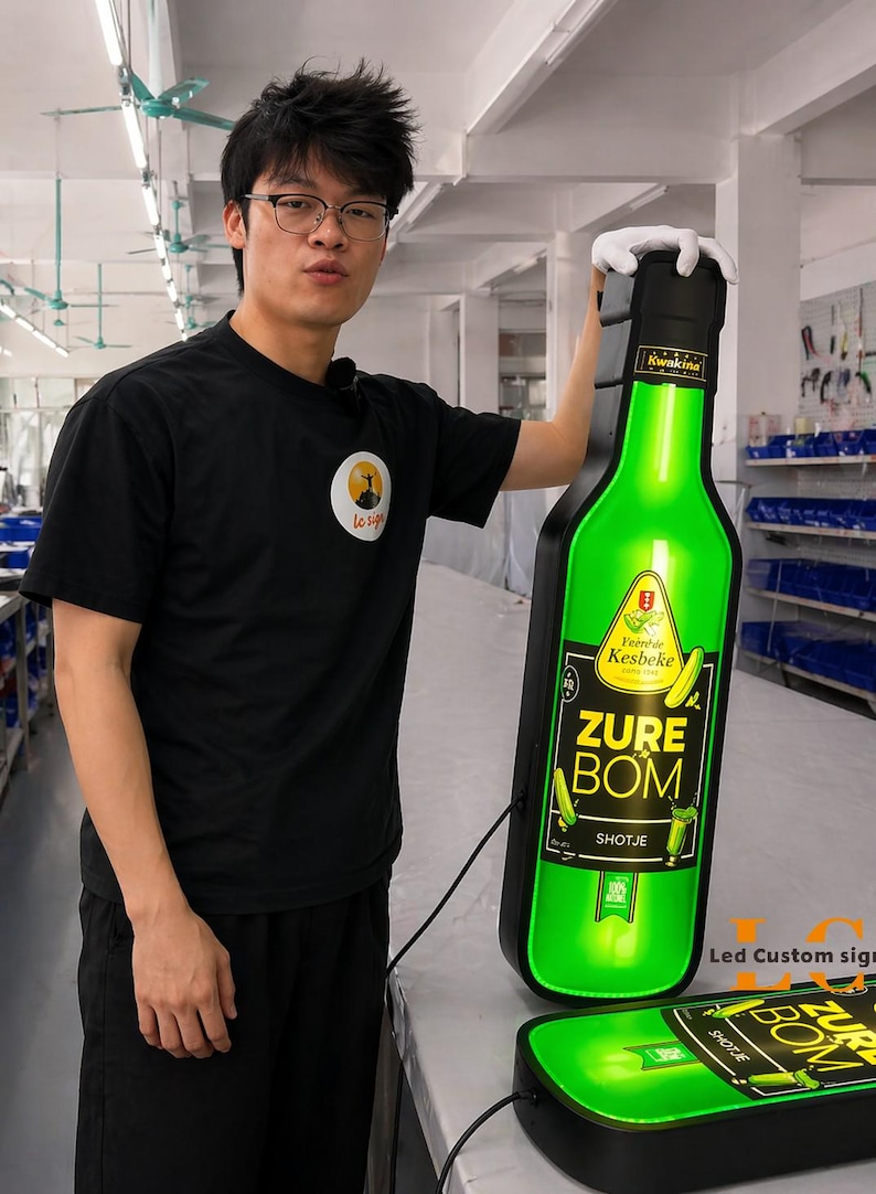 May include: A large, illuminated green bottle-shaped sign with the text "ZURE BOM SHOTJE" and "Veere de Kesbeke" is held by a person. The sign is black-bordered and has a matching base. The person is wearing a black t-shirt.