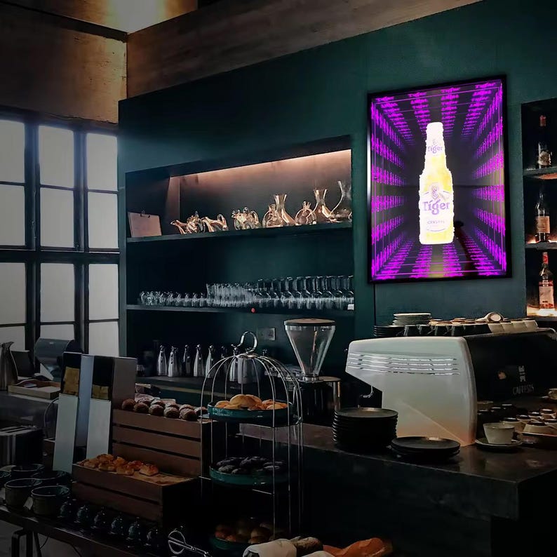 May include: A rectangular wall sign featuring a glowing yellow Tiger beer bottle centered against a vibrant purple and black tunnel-like background. The sign is mounted on a dark green wall above a coffee bar area with shelves and glassware.