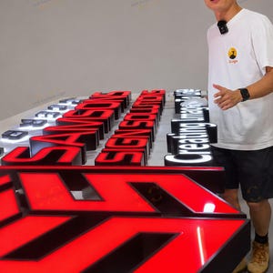 May include: A collection of three-dimensional channel letters with illuminated faces. The signs feature red and white acrylic fronts with black side returns. The text reads SAMSONS SIGN SOLUTIONS and Creating Image in the light, displayed on a flat surface.