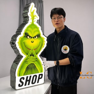 May include: A large, illuminated sign featuring the Grinch's face, with the word "SHOP" at the bottom. The sign has a green and white design with a silver border. The sign is lit up and is being held by a person.