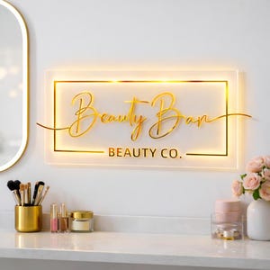 Może przedstawiać: Złoty, podświetlany szyld z napisem "Beauty Bar" i "BEAUTY CO." poniżej. Szyld jest prostokątny ze złotą obw&oacute;dką i jest zamontowany na białej ścianie. Pędzle do makijażu, kosmetyki i kwiaty znajdują się na białej powierzchni.