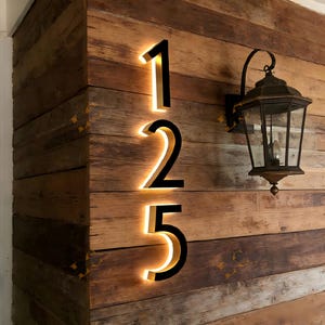 May include: Illuminated house numbers 125 in a modern font, mounted on a rustic wooden wall. The numbers are black with a warm, golden glow emanating from behind. A vintage-style lantern hangs to the right, adding to the welcoming aesthetic.