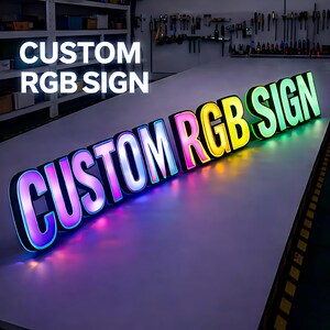 Pode incluir: Uma placa RGB personalizada com as palavras "CUSTOM RGB SIGN" em letras grandes e iluminadas. As letras s&atilde;o contornadas em preto e t&ecirc;m um gradiente de cores, incluindo rosa, azul, amarelo e verde. A placa est&aacute; em uma superf&iacute;cie branca.