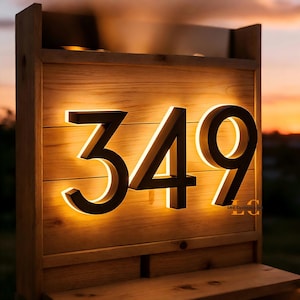 May include: Wooden address sign displaying the numbers 349 in a modern font. The numbers are illuminated with a warm, inviting glow, set against a backdrop of natural wood. The sign is mounted on a wooden structure, perfect for outdoor use.