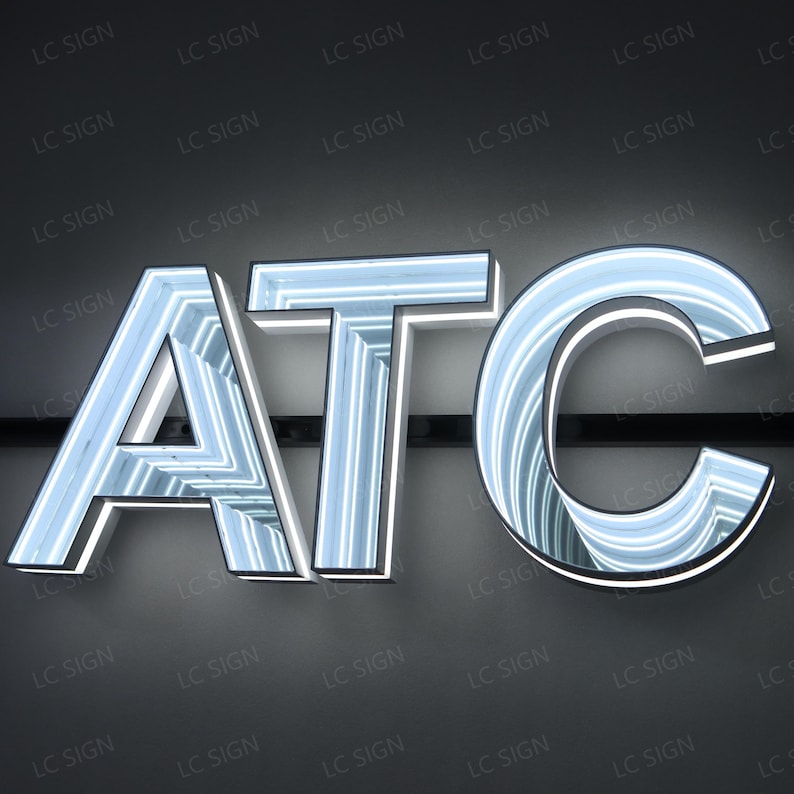 May include: Illuminated sign spelling out "ATC" in a modern font. The letters are white with a light blue glow, creating a 3D effect. The sign is mounted on a dark gray background.