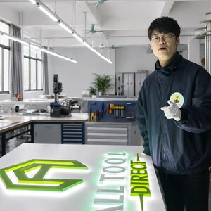 May include: A lit sign with the text "ALL TOOL DIRECT" in white and green. The sign is on a white surface in a workshop setting. A person in a blue jacket and glasses is pointing at the sign.