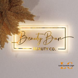 May include: A rectangular LED sign with the words "Beauty Bar" in gold script, and "BEAUTY CO." below. The sign is illuminated with a warm light and framed in gold. The sign is mounted on a white wall.