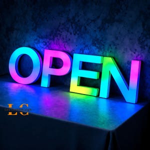 May include: A glowing LED sign spelling "OPEN" in large, block letters. The letters transition through a spectrum of colors, including blue, pink, yellow, and green. The sign is on a reflective surface, with the text "Led custom sign" in the bottom left corner.