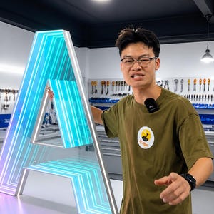 May include: A large, illuminated letter "A" with a gradient of blue and purple lights. The letter is outlined in white and is being held by a person wearing a green t-shirt. The t-shirt has a logo on the front.