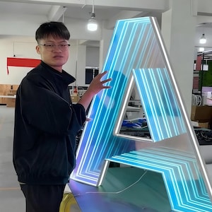 May include: A large, illuminated letter "A" with a white frame and blue neon lights. The letter is standing upright, with a person standing next to it. The neon lights create a striking visual effect, making it suitable for signage or decorative purposes.