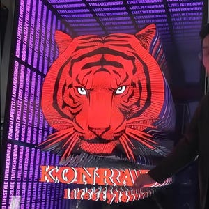 May include: A rectangular poster featuring a large, stylized red tiger head at the center. The background consists of repeating text that reads LIVELIKEKONRAD and KONRAD LIFESTYLE in white. The word KONRAD appears in large orange letters below the tiger.