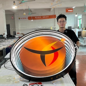 May include: A large circular LED sign featuring a black frame and a bright orange and red center with a stylized black graphic. The sign has a glowing white light ring around the perimeter and is displayed in a workshop setting with visible wiring.