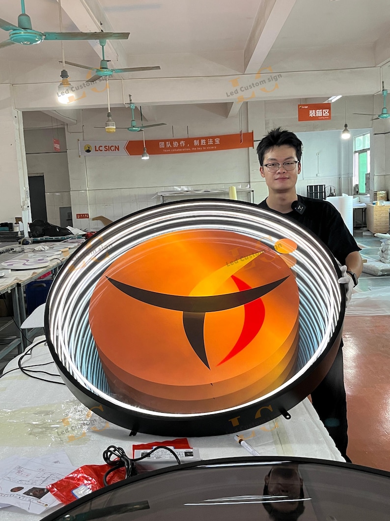 May include: A large circular LED sign featuring a black frame and a bright orange and red center with a stylized black graphic. The sign has a glowing white light ring around the perimeter and is displayed in a workshop setting with visible wiring.