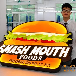 May include: A large, illuminated sign in the shape of a burger. The sign reads "SMASH MOUTH FOODS" with the tagline "YOUR BURGER, YOUR WAY, EST. 2023." The burger design features orange, green, and red colors, with a black border.