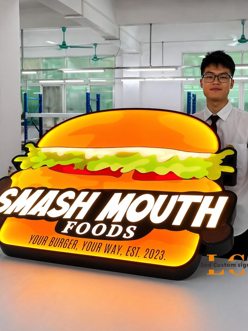 May include: A large, illuminated sign in the shape of a burger. The sign reads "SMASH MOUTH FOODS" with the tagline "YOUR BURGER, YOUR WAY, EST. 2023." The burger design features orange, green, and red colors, with a black border.