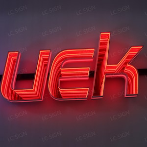 May include: A red neon sign displaying the letters UEK. The sign has a layered design with bright red illumination, creating a striking visual effect. The background is a neutral gray, and the sign is labeled with "LC SIGN".