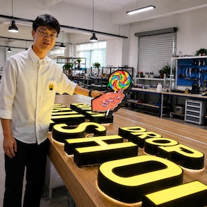 May include: A person holding a colorful lollipop-shaped sign with a hand illustration, positioned next to large, illuminated yellow block letters on a wooden table that spell out the words DROP SHOP in a workshop environment.