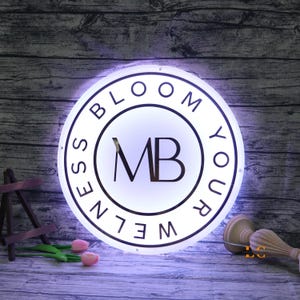 May include: A round, illuminated sign with the words "BLOOM YOUR WELLNESS" in a circular design. The center features the letters "MB" in a bold font. The sign is white with black lettering and is lit from behind.