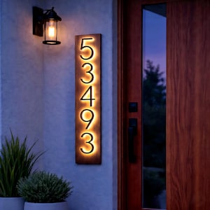May include: A rectangular wooden address sign with the numbers 53493 illuminated in a warm glow. The sign is mounted on a light-colored wall next to a black outdoor light fixture. Green plants are in the foreground.