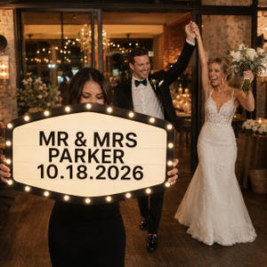 May include: A black and white marquee sign with a pointed oval shape, illuminated by small round bulbs along the perimeter. The sign displays the black text MR & MRS PARKER and the date 10.18.2026 in a clean, bold, sans-serif font style.
