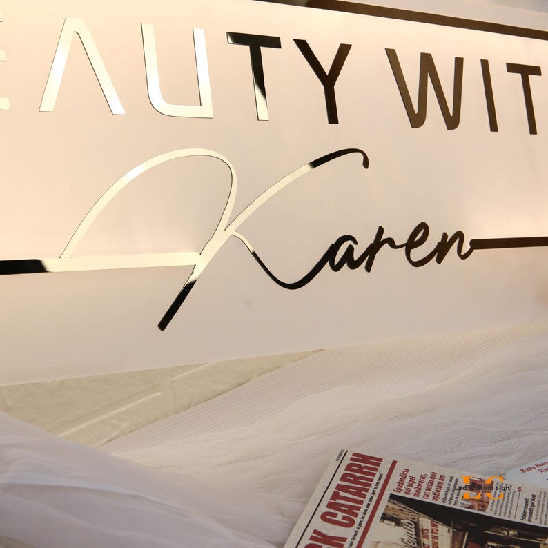 May include: A sign with the words "BEAUTY WITH" in silver mirrored letters and the name "Karen" in a black cursive font. The sign is mounted on a white background.